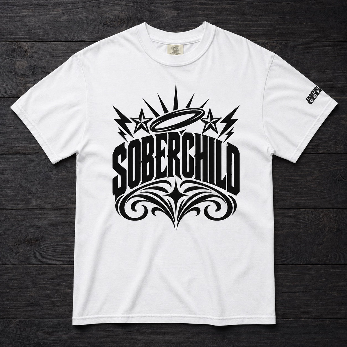 A white short-sleeve crewneck t-shirt with a large black graphic on the front reading 'SOBERCHILD' in stylized lettering, featuring stars, lightning bolts, and ornate flourishes. The shirt is laid flat on a dark wood plank background.