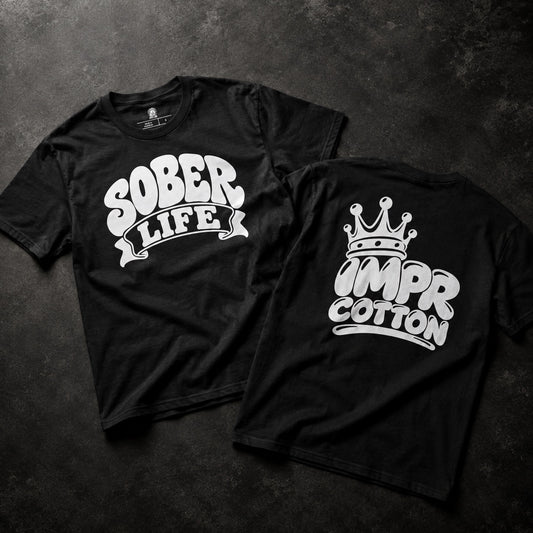 Two black short-sleeve T-shirts laid flat on a dark textured surface, showing the front and back designs. The front shirt has a white graphic reading “SOBER LIFE,” and the back shirt has a white graphic with a crown above stylized text that appears to read “IMPR” and “COTTON.