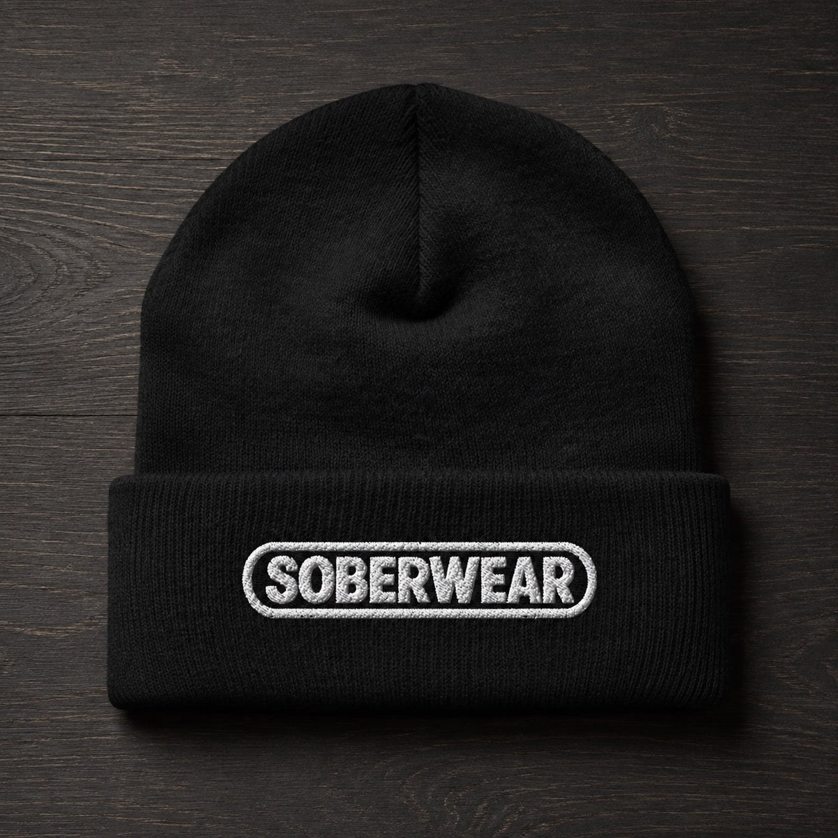 A black knit beanie with a folded cuff featuring a white embroidered oval logo that reads 'SOBERWEAR', displayed flat on a dark wood background.