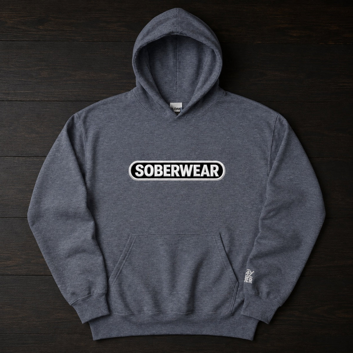 A dark heather gray pullover hoodie with a white and black 'SOBERWEAR' logo on the chest, laid flat on a dark wood background.