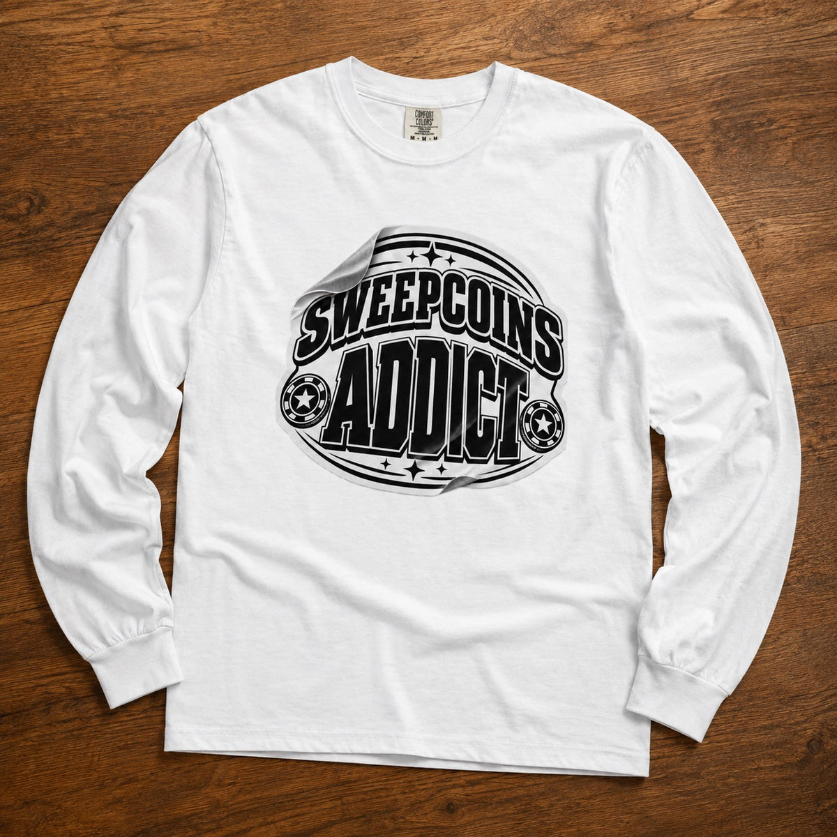 A white long-sleeve t-shirt with a black graphic print reading 'SWEEPCOINS ADDICT' on the front, laid flat on a brown wooden surface.