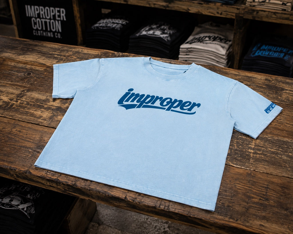 A light blue crewneck t-shirt with a dark blue 'improper' logo across the chest, displayed on a rustic wooden table in a retail store setting.