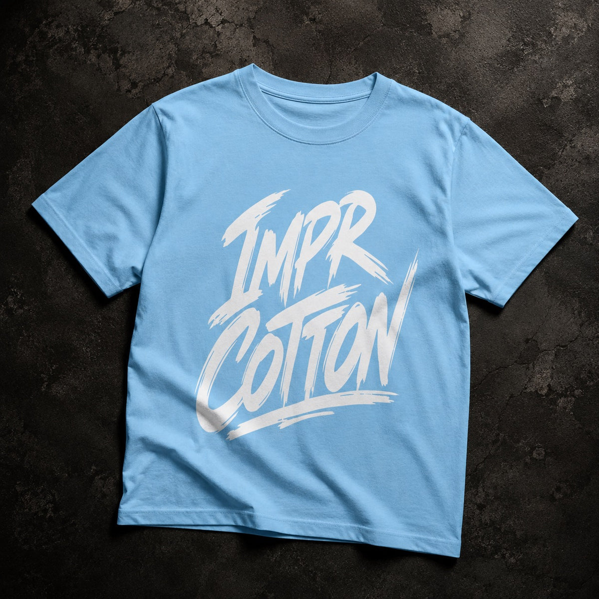 A light blue crewneck t-shirt with a white brush-stroke style graphic reading 'IMPR COTTON' on the front, laid flat on a dark textured background.