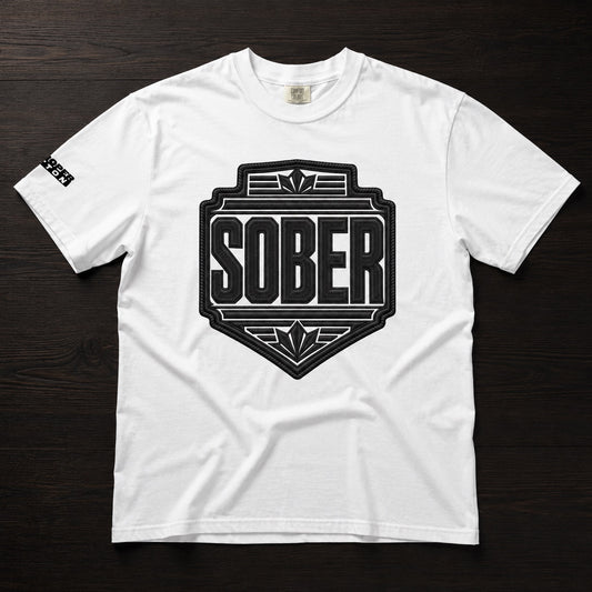 A white short-sleeve crewneck t-shirt with a large black shield graphic on the chest reading 'SOBER' in bold letters, laid flat on a dark wood background.