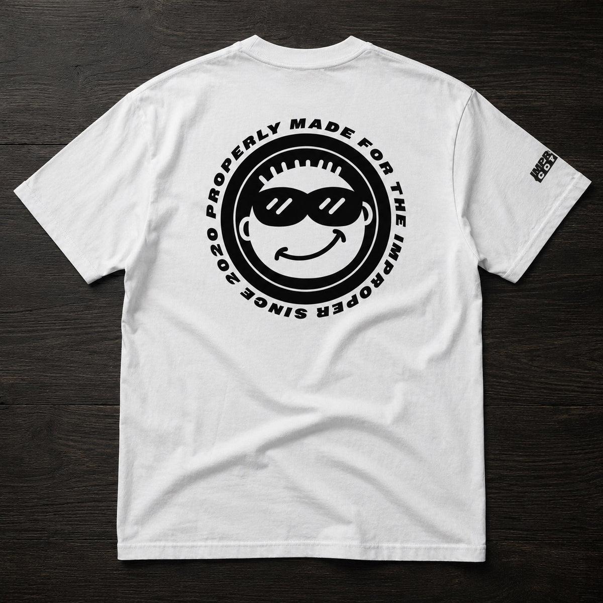 A white short-sleeve t-shirt laid flat on a dark wood background, featuring a large black circular graphic on the back with a smiley face wearing sunglasses and the text 'PROPERLY MADE FOR THE IMPROPER SINCE 2020'. A small black logo is printed on the left sleeve.