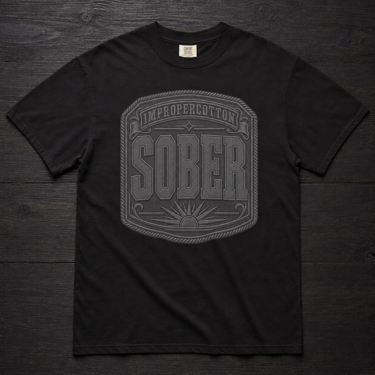 A black short-sleeve crewneck t-shirt laid flat on a dark wood background, featuring a large gray graphic on the front with the words 'IMPROPERCOTTON SOBER' inside a decorative border.