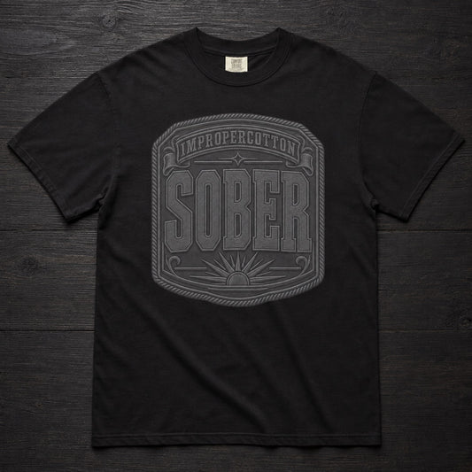A black short-sleeve crewneck t-shirt laid flat on a dark wood background, featuring a large gray graphic on the front with the words 'IMPROPERCOTTON SOBER' inside a decorative border.