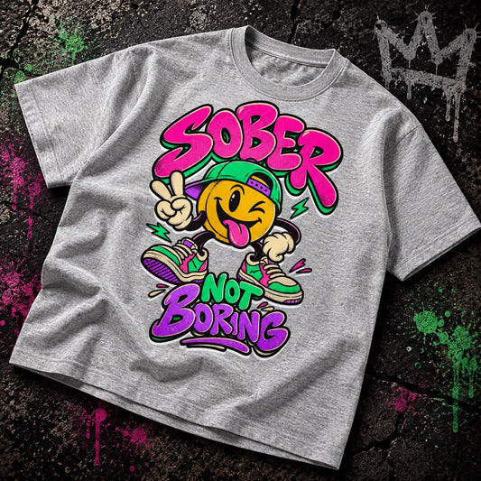 A heather gray graphic t-shirt laid flat on a dark, graffiti-splattered background. The graphic features a yellow smiley face wearing a green cap and colorful sneakers, with the text 'SOBER NOT BORING' in vibrant pink, purple, and green bubble lettering.