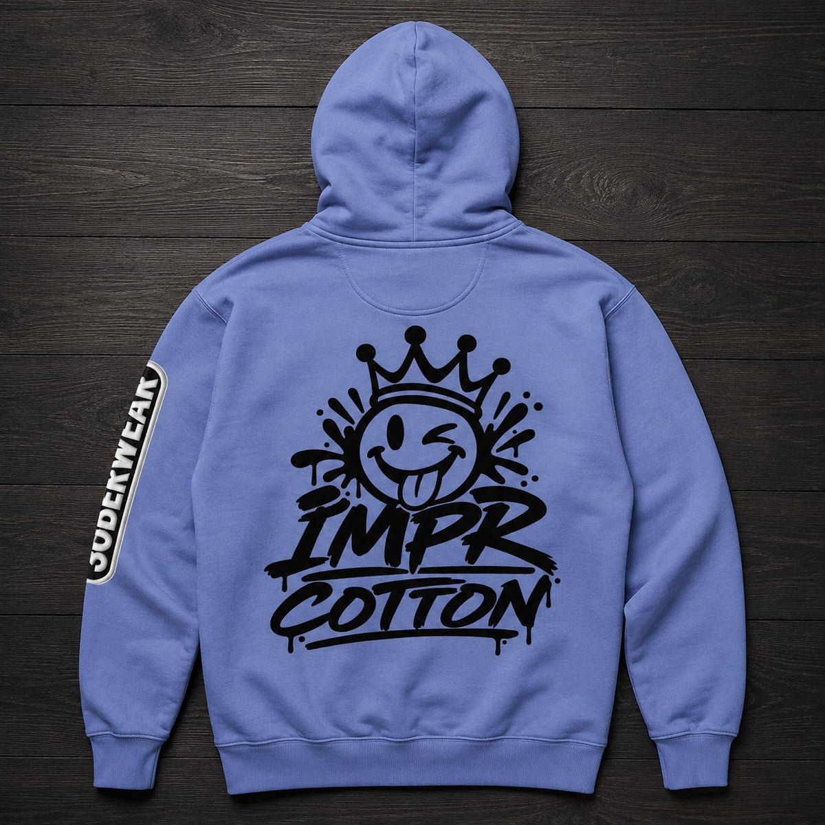 A light blue hoodie shown from the back, laid flat on a dark wood background. The back features a large black graphic of a winking smiley face under a crown with the text 'IMPR COTTON'.