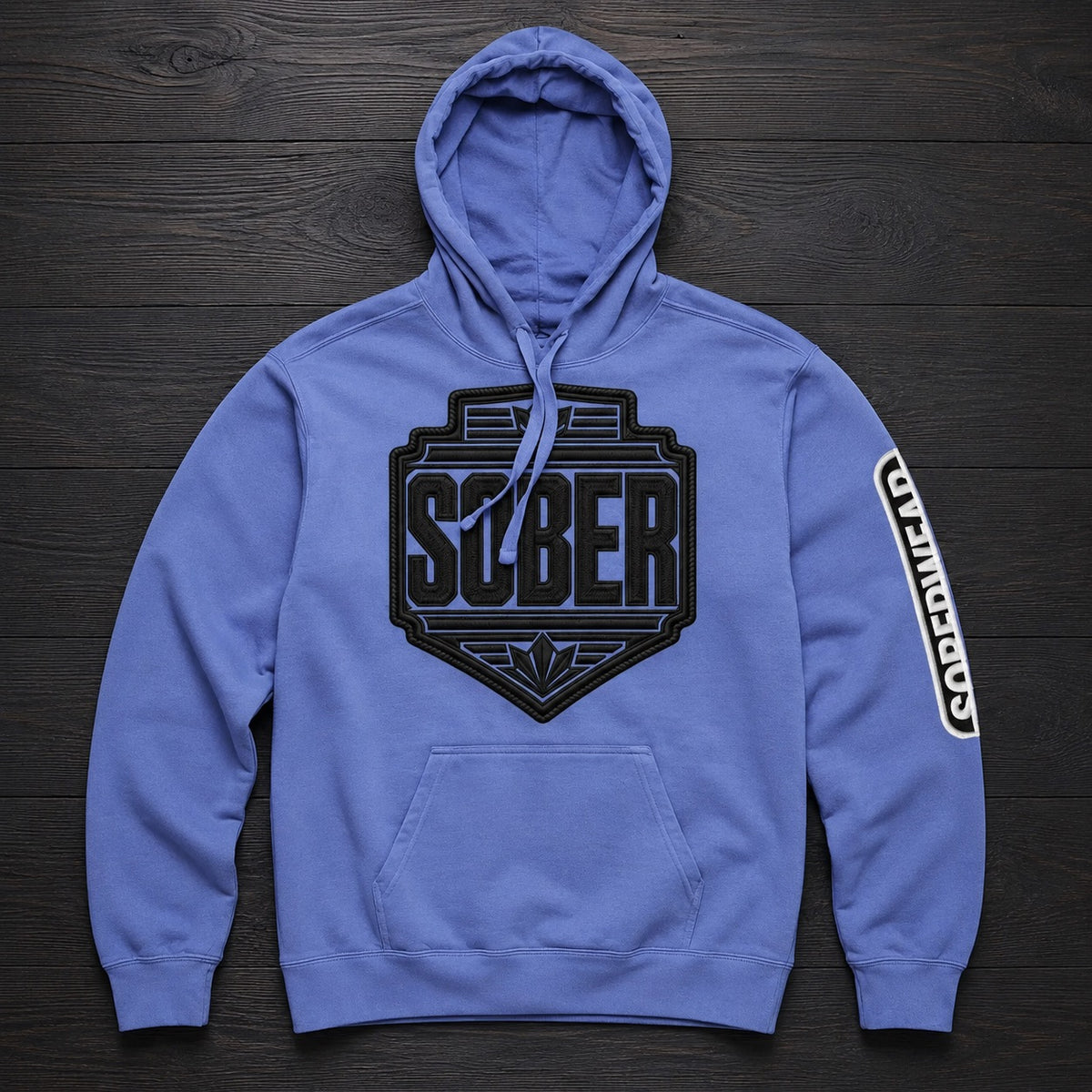 A blue pullover hoodie laid flat on a dark wood background, featuring a large black 'SOBER' shield logo on the chest and a vertical white-and-black 'SOBERWEAR' graphic on the right sleeve.