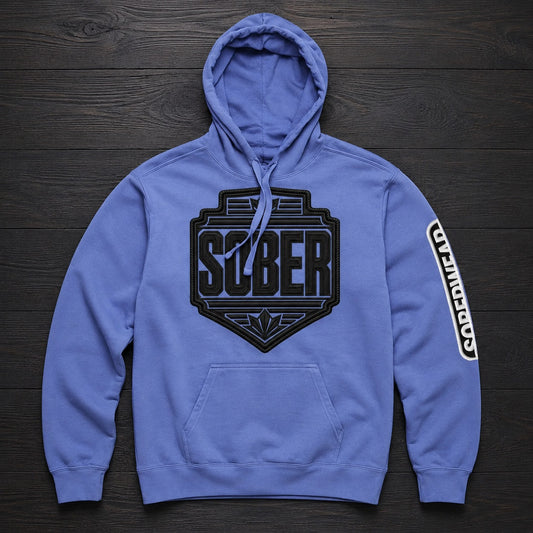 A blue pullover hoodie laid flat on a dark wood background, featuring a large black 'SOBER' shield logo on the chest and a vertical white-and-black 'SOBERWEAR' graphic on the right sleeve.