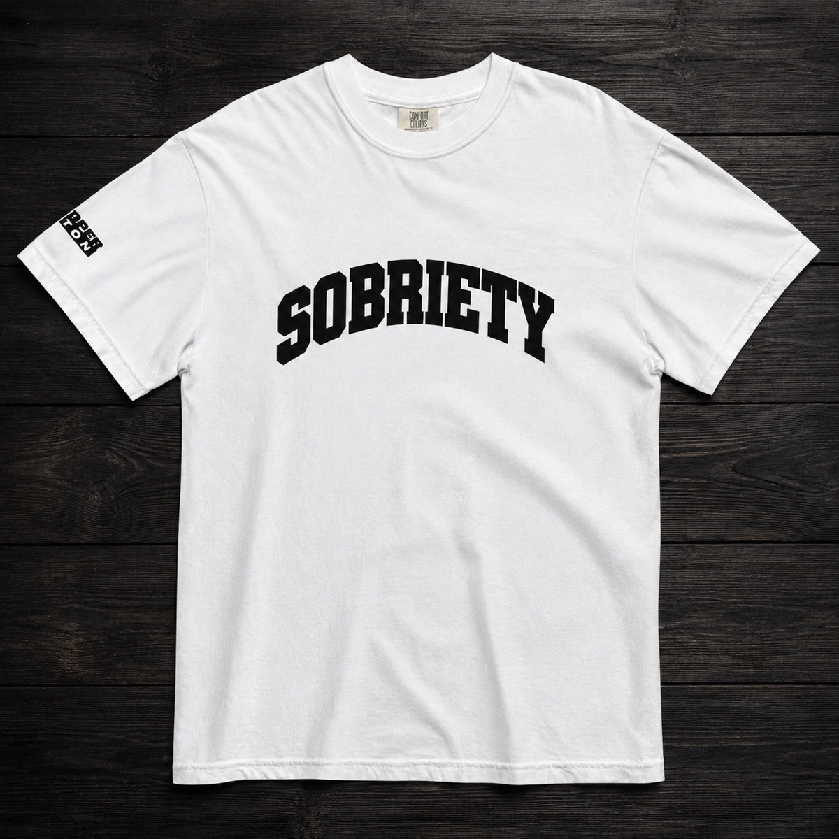 A white short-sleeve crewneck t-shirt with 'SOBRIETY' printed in black arched varsity letters across the chest, laid flat on a dark wood plank background.