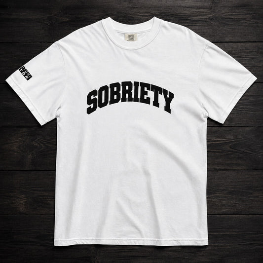 A white short-sleeve crewneck t-shirt with 'SOBRIETY' printed in black arched varsity letters across the chest, laid flat on a dark wood plank background.