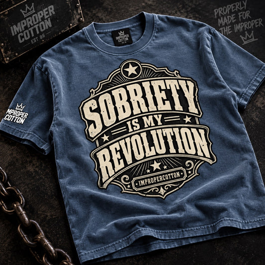 A blue graphic t-shirt laid flat on a dark textured background with vintage-style props, including a chain and a sign reading 'Improper Cotton.' The shirt features a large cream graphic that reads 'Sobriety is my revolution' with small crown logos and 'ImproperCotton' branding visible on the neck label and sleeve.