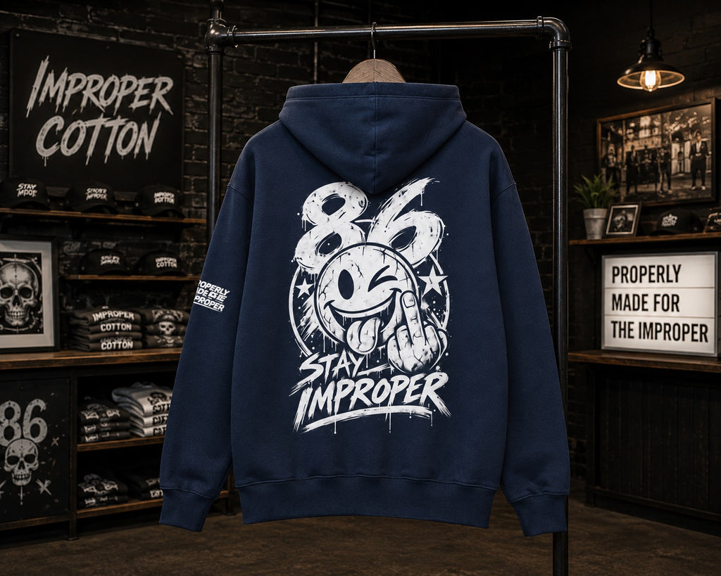 A navy blue hoodie displayed on a clothing rack in a dark retail store setting, showing the back view with a large white graphic print featuring the number 86, a smiley face with its tongue sticking out, and the words 'Stay Improper'.