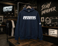A navy blue hoodie with a bold white 'IMPROPER COTTON' logo across the chest, displayed on a hanger on a clothing rack inside a branded retail store.