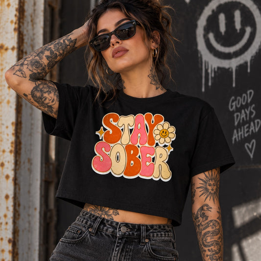 A woman wearing a black crop top t-shirt with a retro-style 'STAY SOBER' graphic in pink, orange, and yellow, paired with dark gray jeans. She is posing in front of a weathered metal wall and a black wall with graffiti.