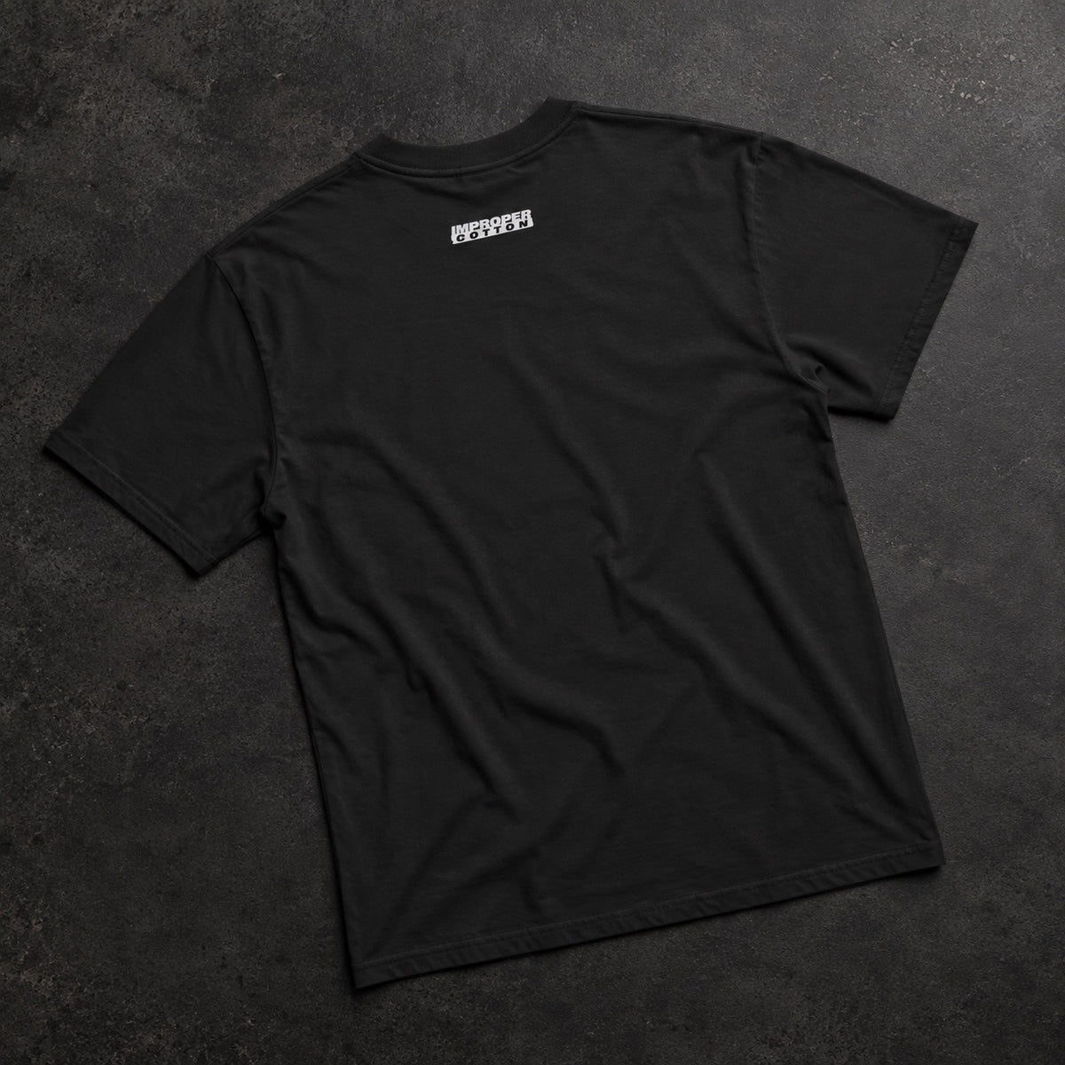 A black short-sleeve t-shirt laid flat on a dark textured surface, viewed from the back. It features a small white rectangular graphic printed on the upper center back.