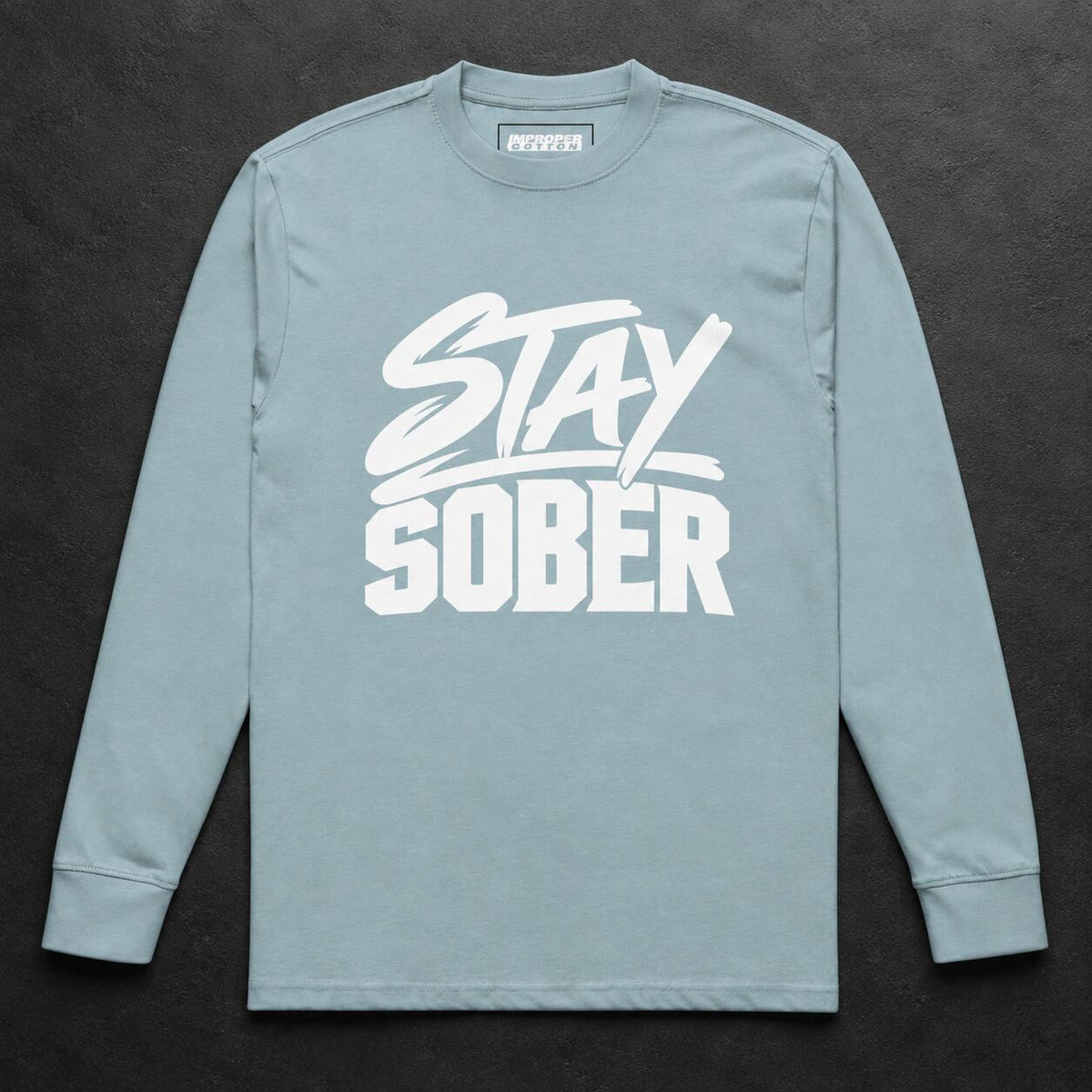 A light blue long-sleeve t-shirt with a white graphic print reading 'STAY SOBER' on the front, laid flat against a dark textured background.