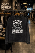 A black acid-wash long-sleeve t-shirt with a white graphic print reading 'STAY IMPROPER' and a winking face with a middle finger, hanging on a clothing rack inside a retail store.