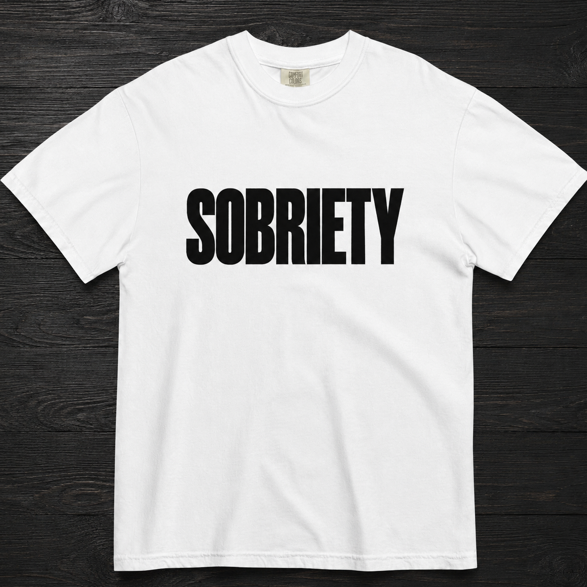 A white short-sleeve crewneck t-shirt with the word 'SOBRIETY' printed in bold black capital letters across the chest, laid flat on a dark wood background.
