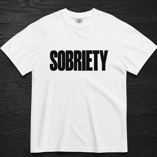 A white short-sleeve crewneck t-shirt with the word 'SOBRIETY' printed in bold black capital letters across the chest, laid flat on a dark wood background.