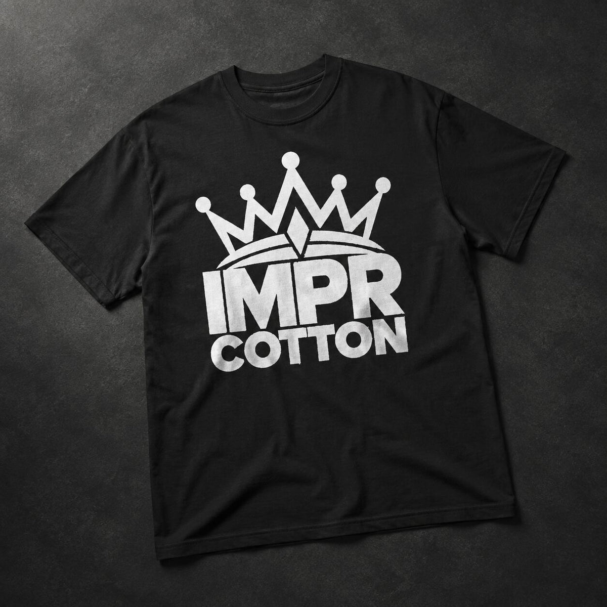 A black short-sleeve t-shirt laid flat on a dark textured background, featuring a white graphic of a crown above the words 'IMPR COTTON' in bold white letters.