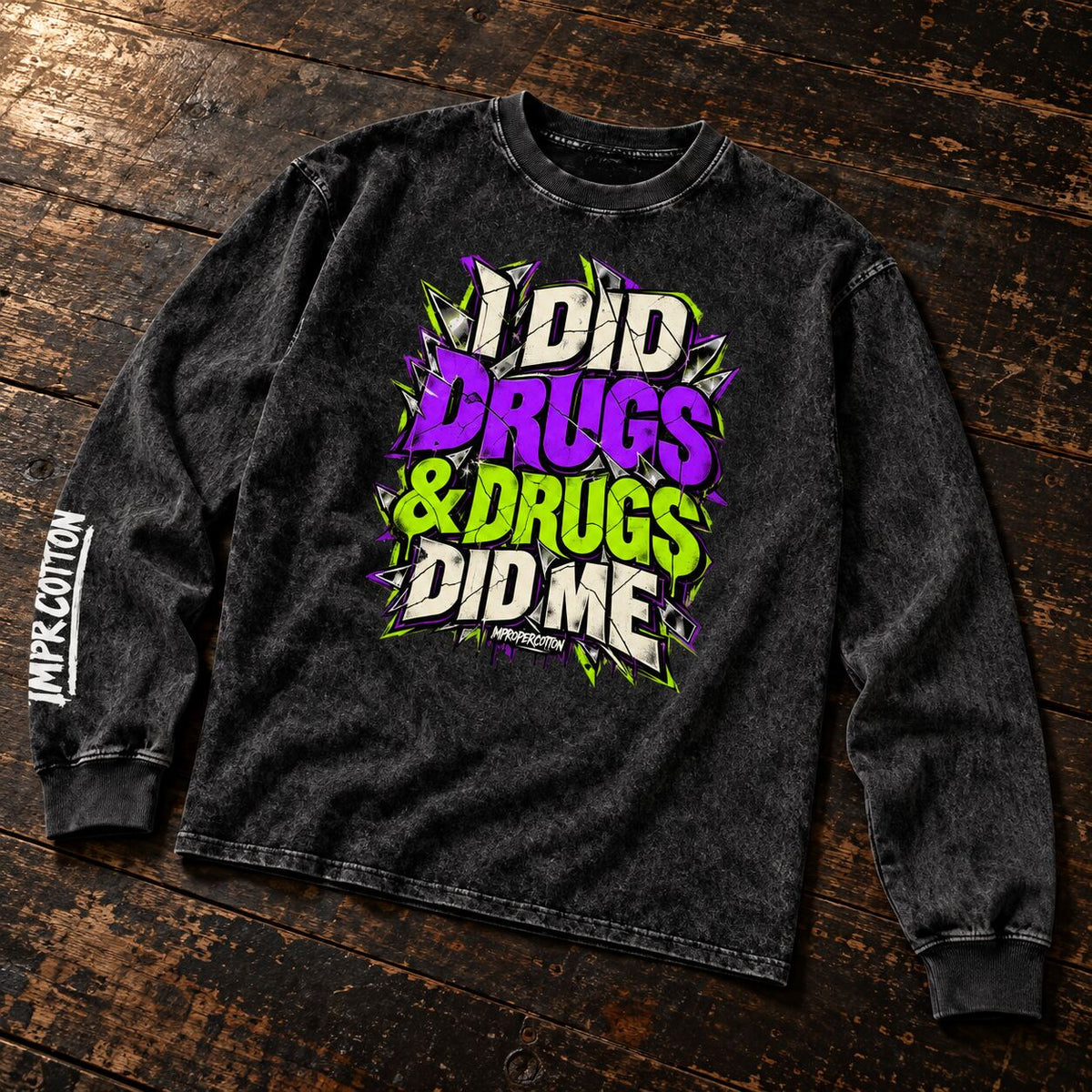 A black acid-wash long-sleeve t-shirt with a colorful graphic reading 'I DID DRUGS & DRUGS DID ME' in purple, green, and white. The shirt is laid flat on a rustic dark wood background, with 'IMPACT COTTON' printed vertically on the left sleeve.