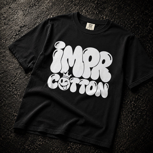 A black short-sleeve t-shirt laid flat on a dark textured surface, featuring a large white graphic print that reads 'IMPR COTTON' with a small crown and smiley face detail.