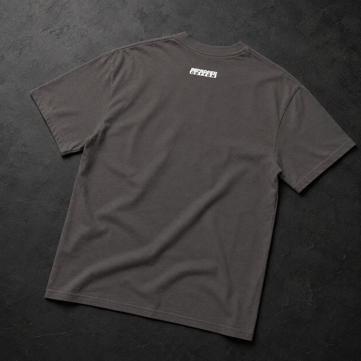 A dark gray crewneck t-shirt laid flat on a dark textured background, featuring a small white rectangular graphic printed on the upper back.