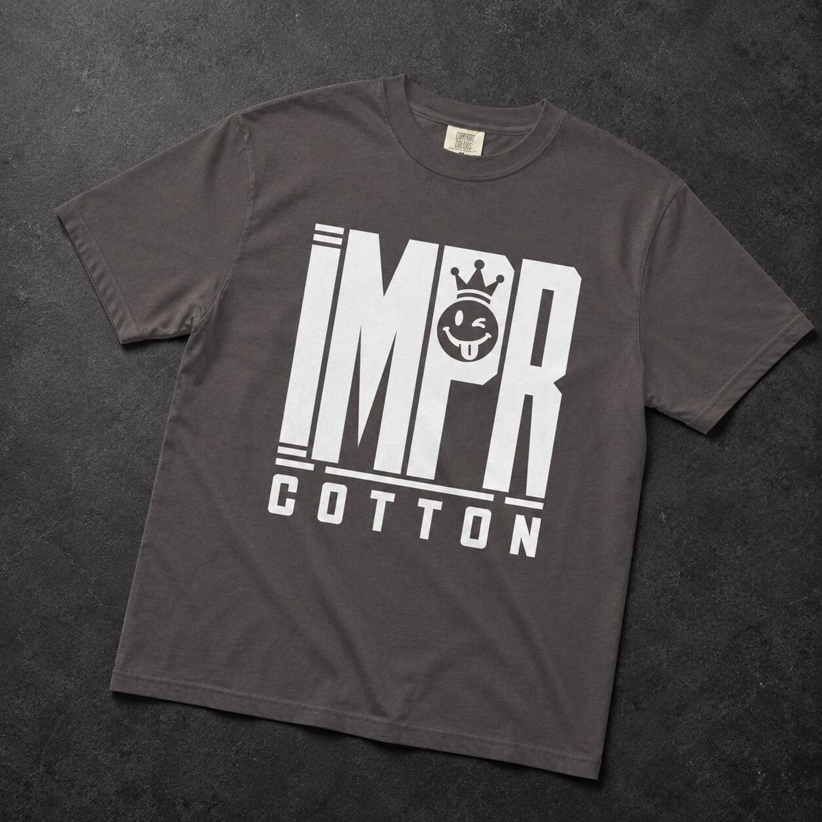 A dark gray crewneck t-shirt with a bold white graphic print reading 'IMPR COTTON' and a crown motif, laid flat on a dark textured background.