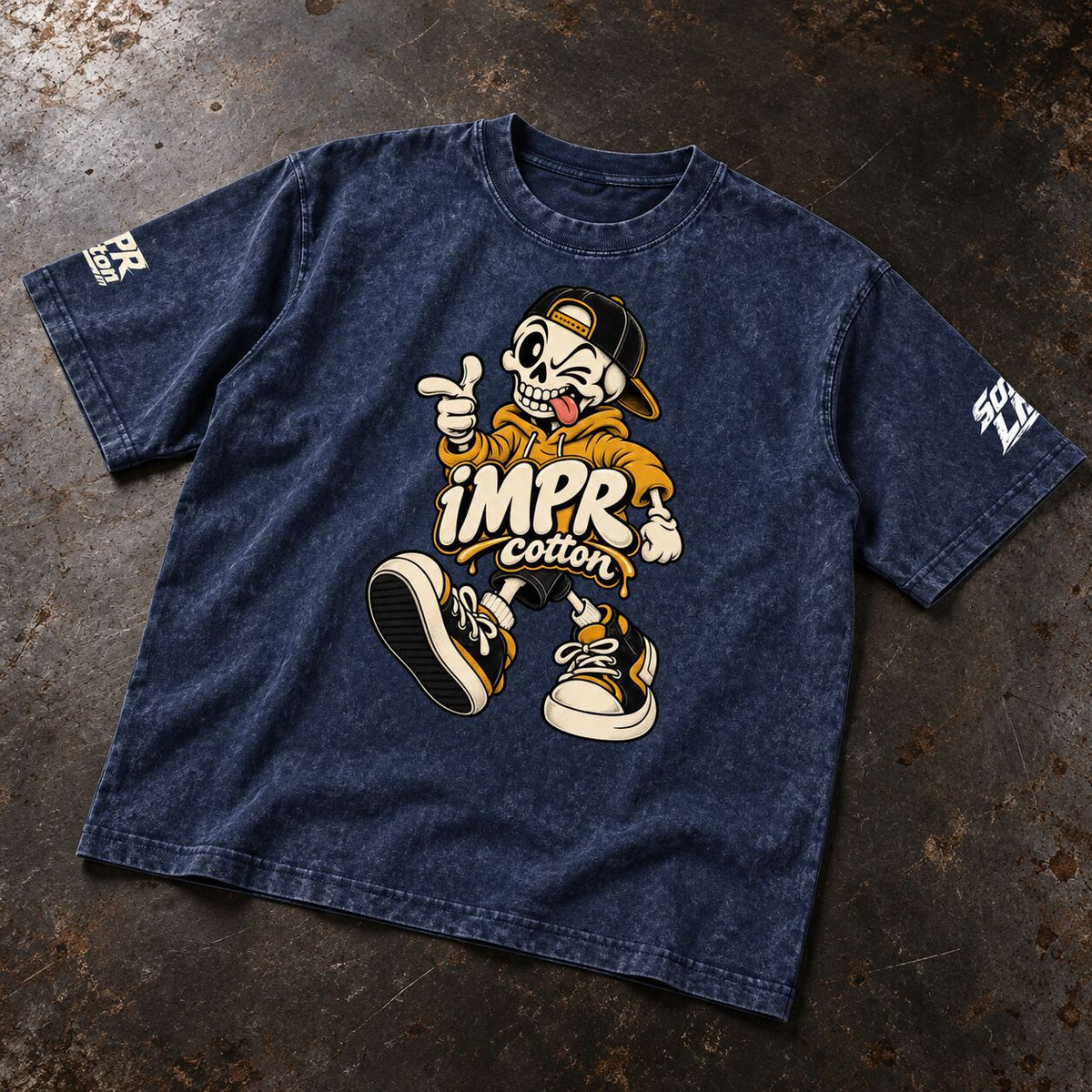 A dark blue acid-wash graphic t-shirt laid flat on a textured dark gray surface. The shirt features a large cartoon graphic of a skeleton in a yellow hoodie and cap with the text 'iMPR cotton' in the center, and white graphic text on both sleeves.