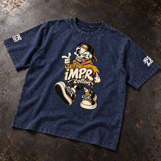 A dark blue acid-wash graphic t-shirt laid flat on a textured dark gray surface. The shirt features a large cartoon graphic of a skeleton in a yellow hoodie and cap with the text 'iMPR cotton' in the center, and white graphic text on both sleeves.