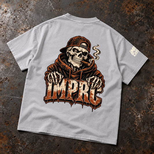 A light gray t-shirt laid flat on a textured, rusty dark gray background. The back of the shirt features a large graphic of a skull wearing a brown hood and cap, smoking a cigar with smoke rising, above the stylized text 'IMPC' in a distressed orange and brown font.