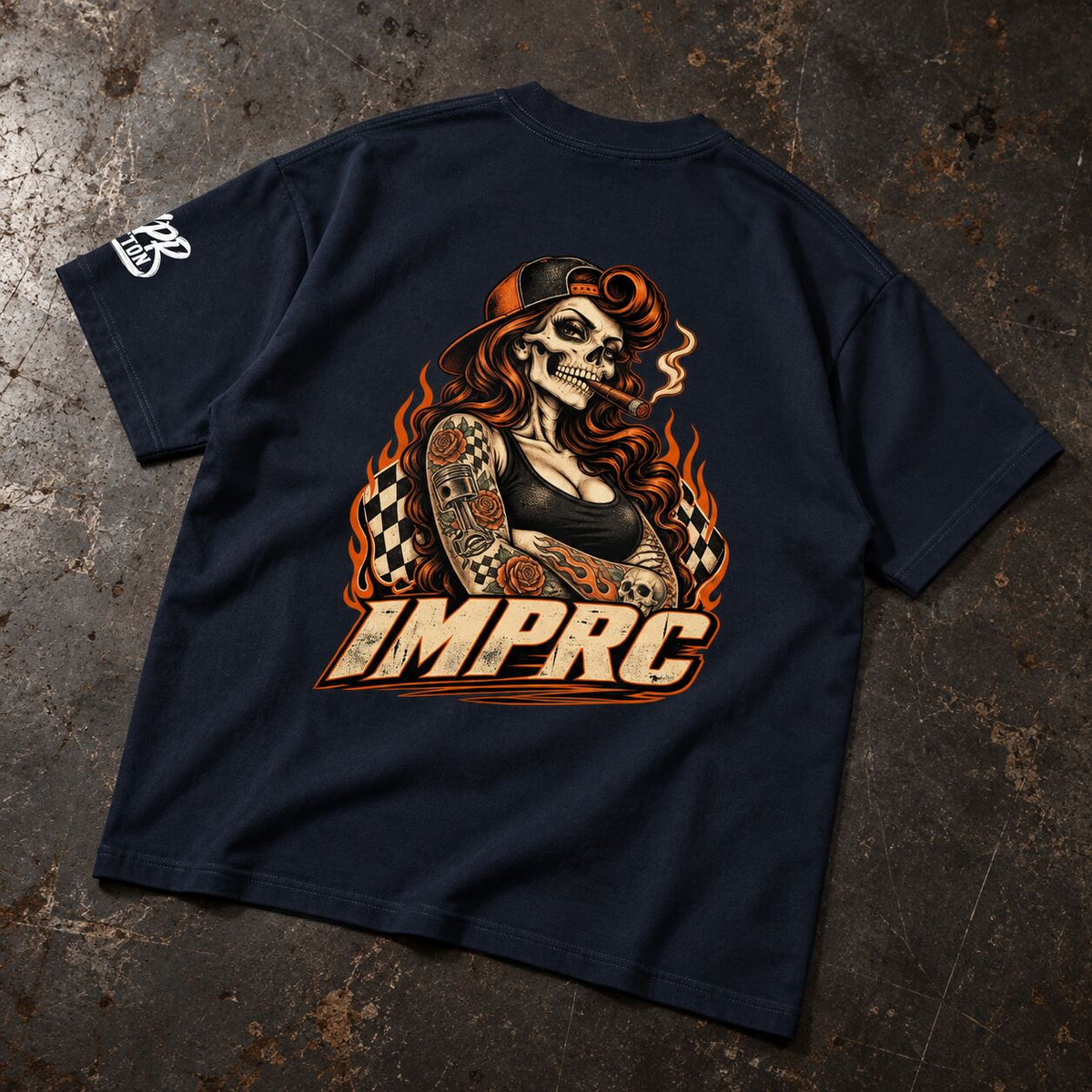A navy blue t-shirt laid flat on a textured, dark metallic surface, showing the back graphic. The graphic features a stylized skeleton woman with long red hair, a smoking cigar, and tattoos, with the text 'IMPRC' in a bold, retro font below.