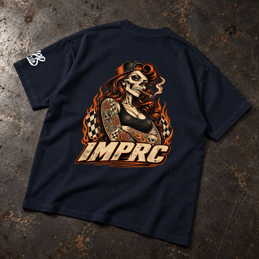 A navy blue t-shirt laid flat on a textured, dark metallic surface, showing the back graphic. The graphic features a stylized skeleton woman with long red hair, a smoking cigar, and tattoos, with the text 'IMPRC' in a bold, retro font below.