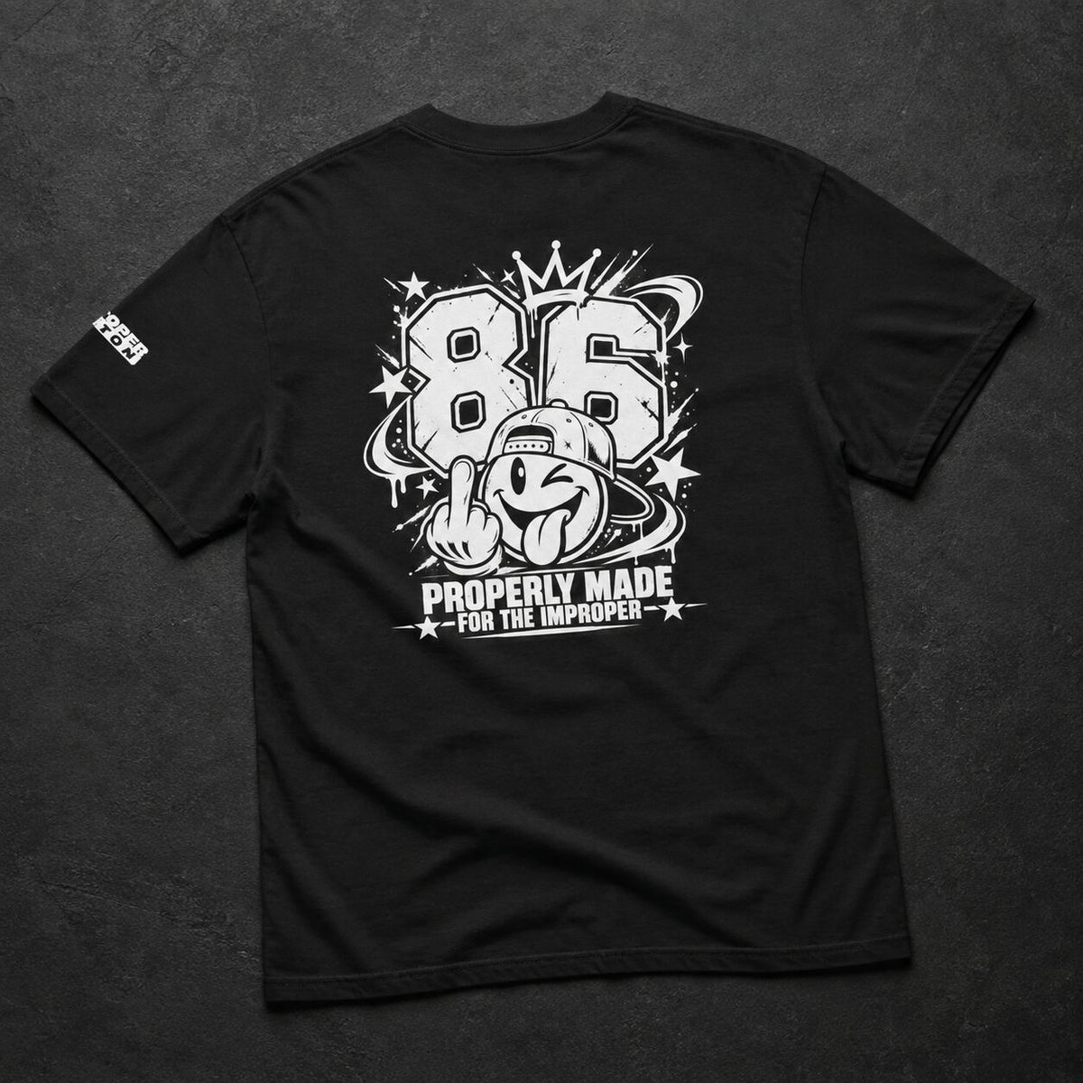 A black short-sleeve t-shirt laid flat on a dark textured background, showing a large white graphic print on the back with the number 86, a cartoon character, stars, and the text 'PROPERLY MADE FOR THE IMPROPER'. A small white logo is visible on the left sleeve.