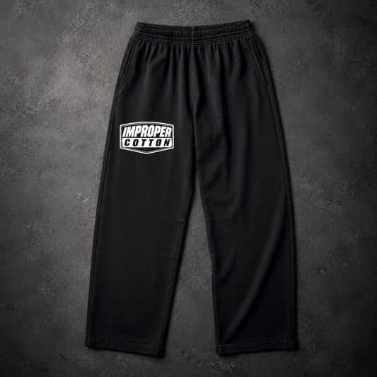 Black sweatpants with an elastic waistband and a white 'IMPROPER COTTON' graphic on the left thigh, displayed flat against a dark textured background.