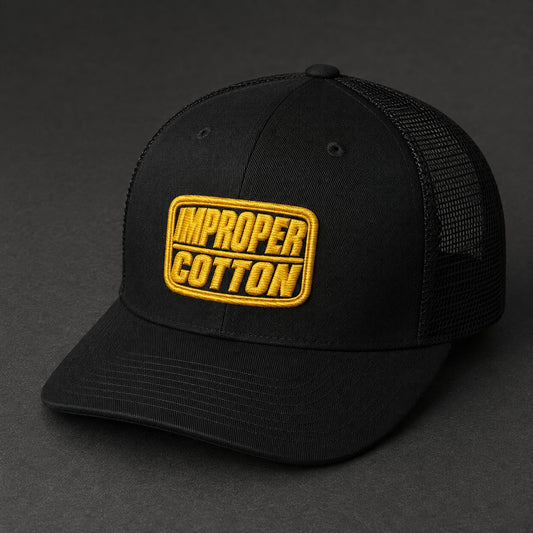 A black trucker-style cap with a yellow embroidered patch reading 'IMPROPER COTTON' on the front, photographed on a dark gray background.