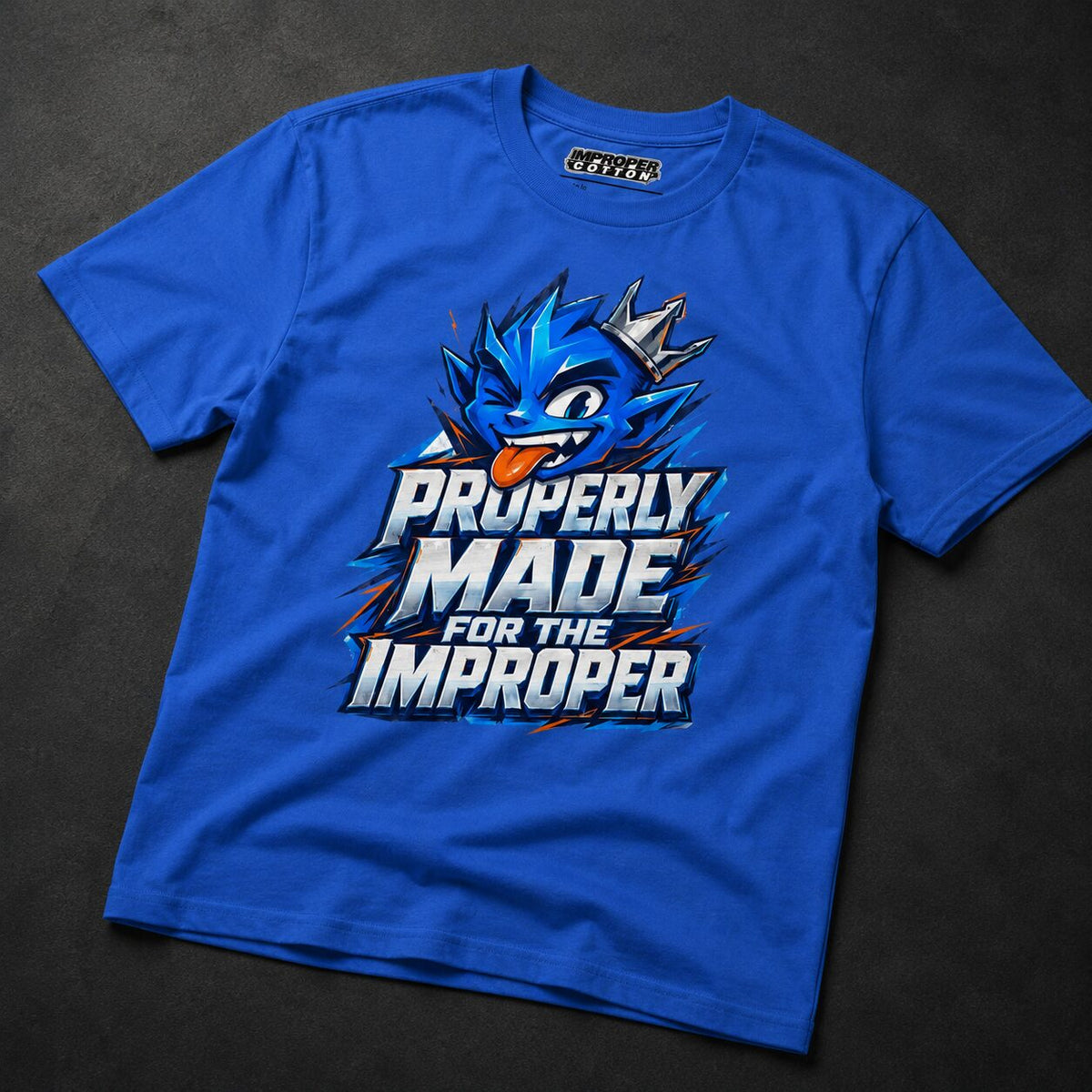 A royal blue short-sleeve t-shirt with a graphic print of a blue cartoon character and the text 'PROPERLY MADE FOR THE IMPROPER' in white, blue, and orange, laid flat on a dark textured background.