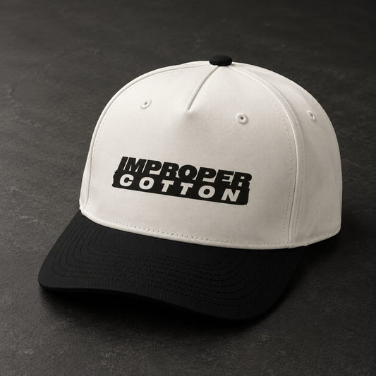 A structured baseball cap with an off-white crown and a black curved brim, featuring the text 'IMPROPER COTTON' in black on the front. The cap is photographed on a dark, textured slate surface.