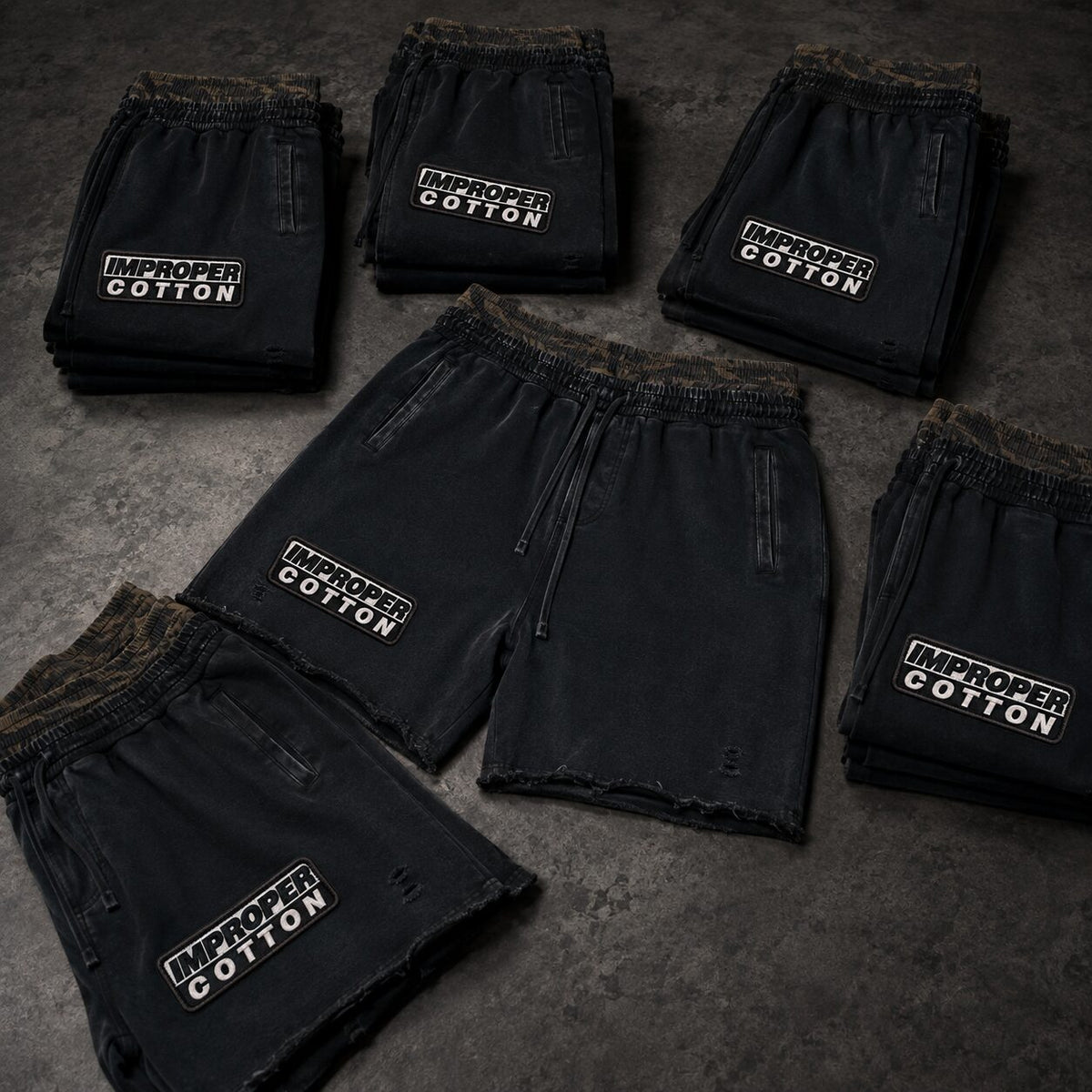 Five pairs of black distressed shorts arranged on a dark textured surface, each with a white embroidered 'IMPROPER COTTON' logo on the left leg.