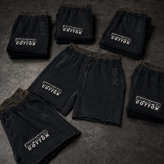 Five pairs of black distressed shorts arranged on a dark textured surface, each with a white embroidered 'IMPROPER COTTON' logo on the left leg.