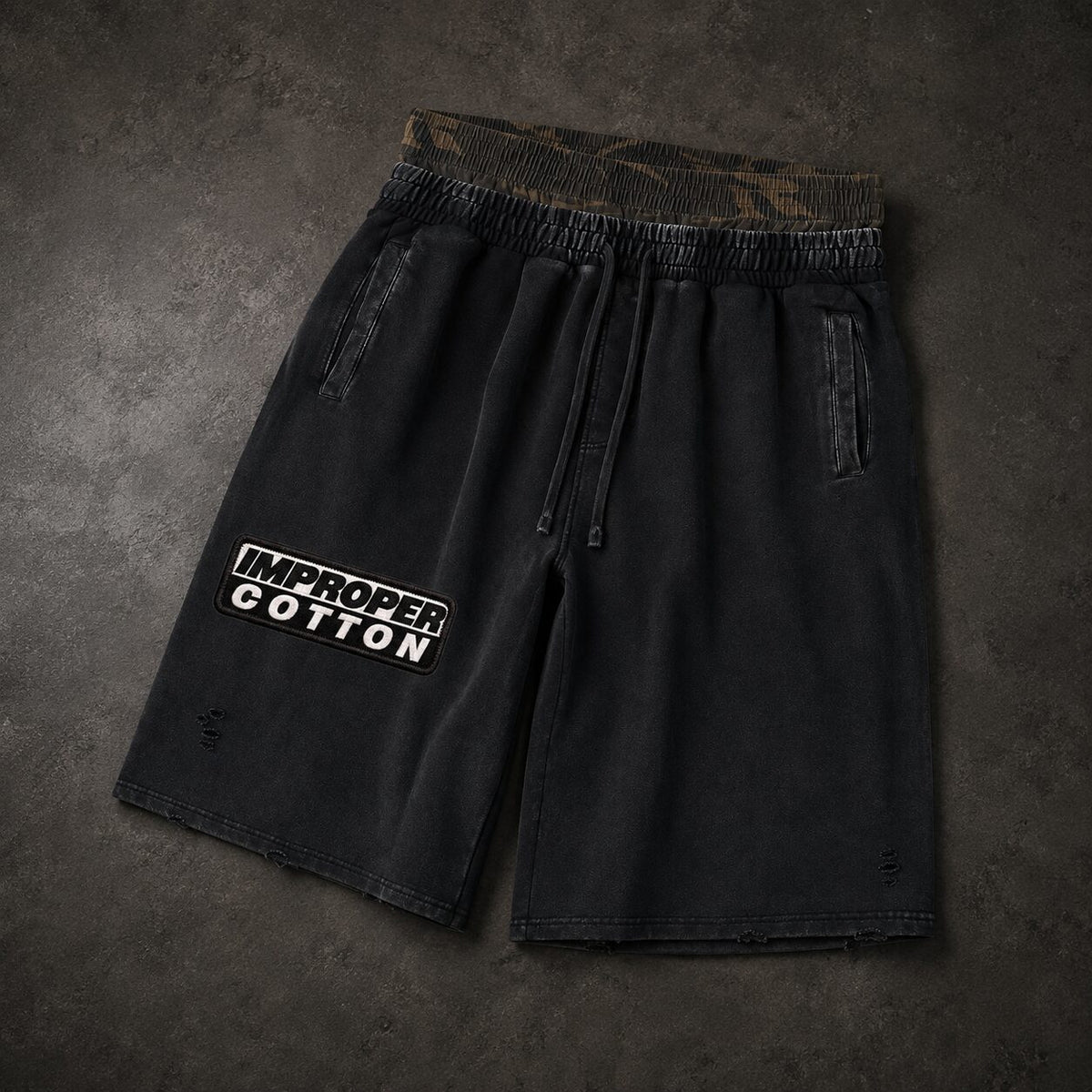 Black acid-wash distressed shorts with a camouflage patterned elastic waistband, featuring a white and black 'IMPROPER COTTON' graphic on the left leg, laid flat on a dark textured background.