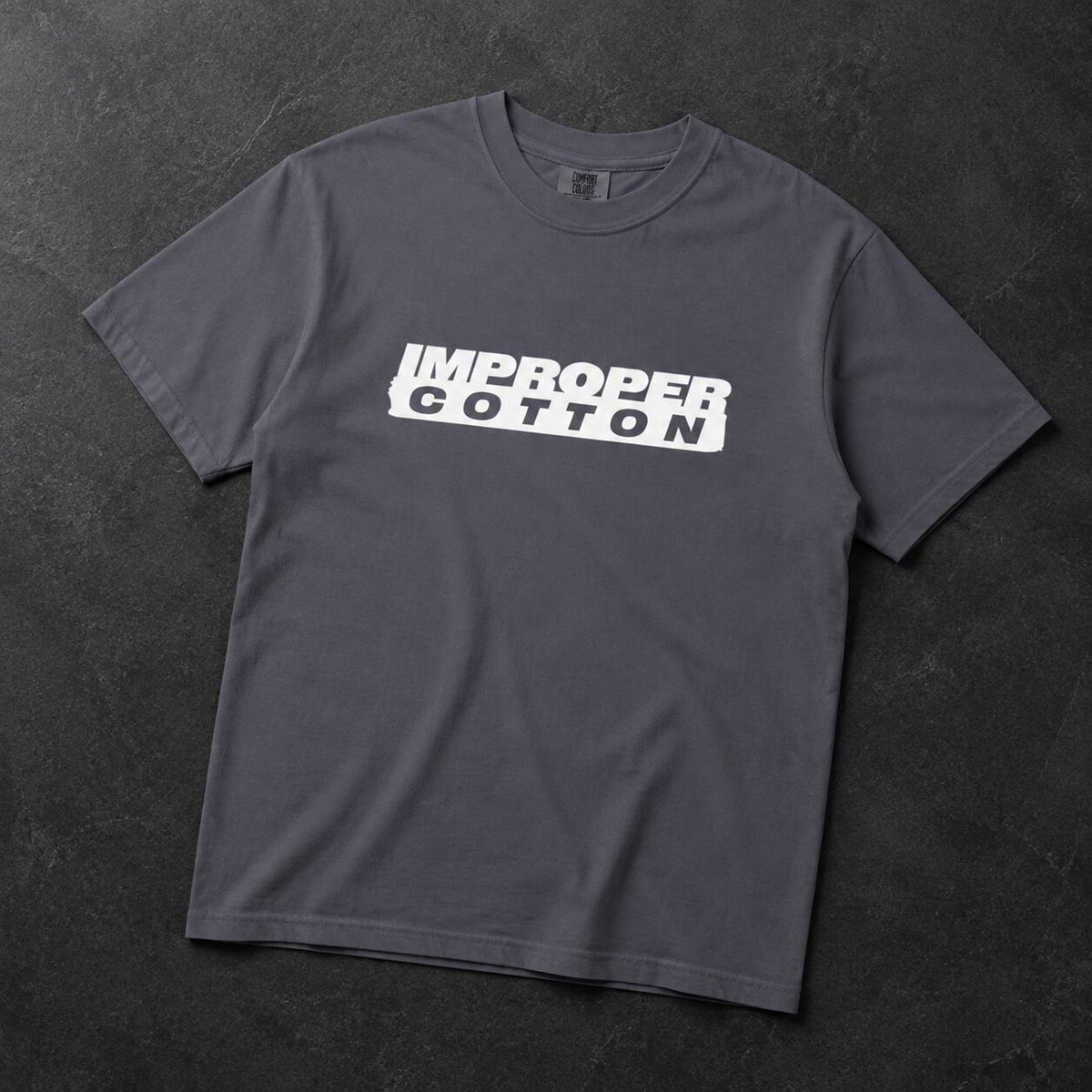 A dark gray crewneck t-shirt with a white graphic print reading 'IMPROPER COTTON' across the chest, laid flat on a dark textured background.
