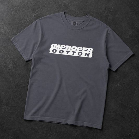 A dark gray crewneck t-shirt with a white graphic print reading 'IMPROPER COTTON' across the chest, laid flat on a dark textured background.