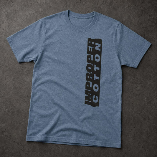 A heather blue crewneck t-shirt laid flat on a textured dark gray surface, featuring a vertical black graphic print on the left chest that reads 'IMPROPER COTTON'.
