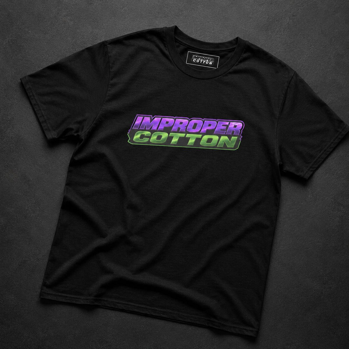 A black short-sleeve t-shirt laid flat on a dark textured background, featuring a colorful 'IMPROPER COTTON' graphic across the chest in purple and green.