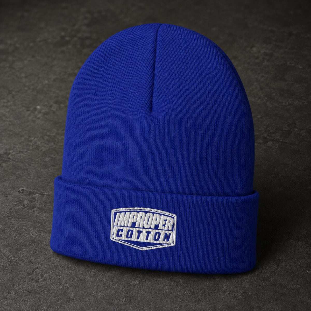 A royal blue ribbed knit beanie hat with a folded cuff, featuring an embroidered 'IMPROPER COTTON' logo patch on the front, set against a dark textured background.