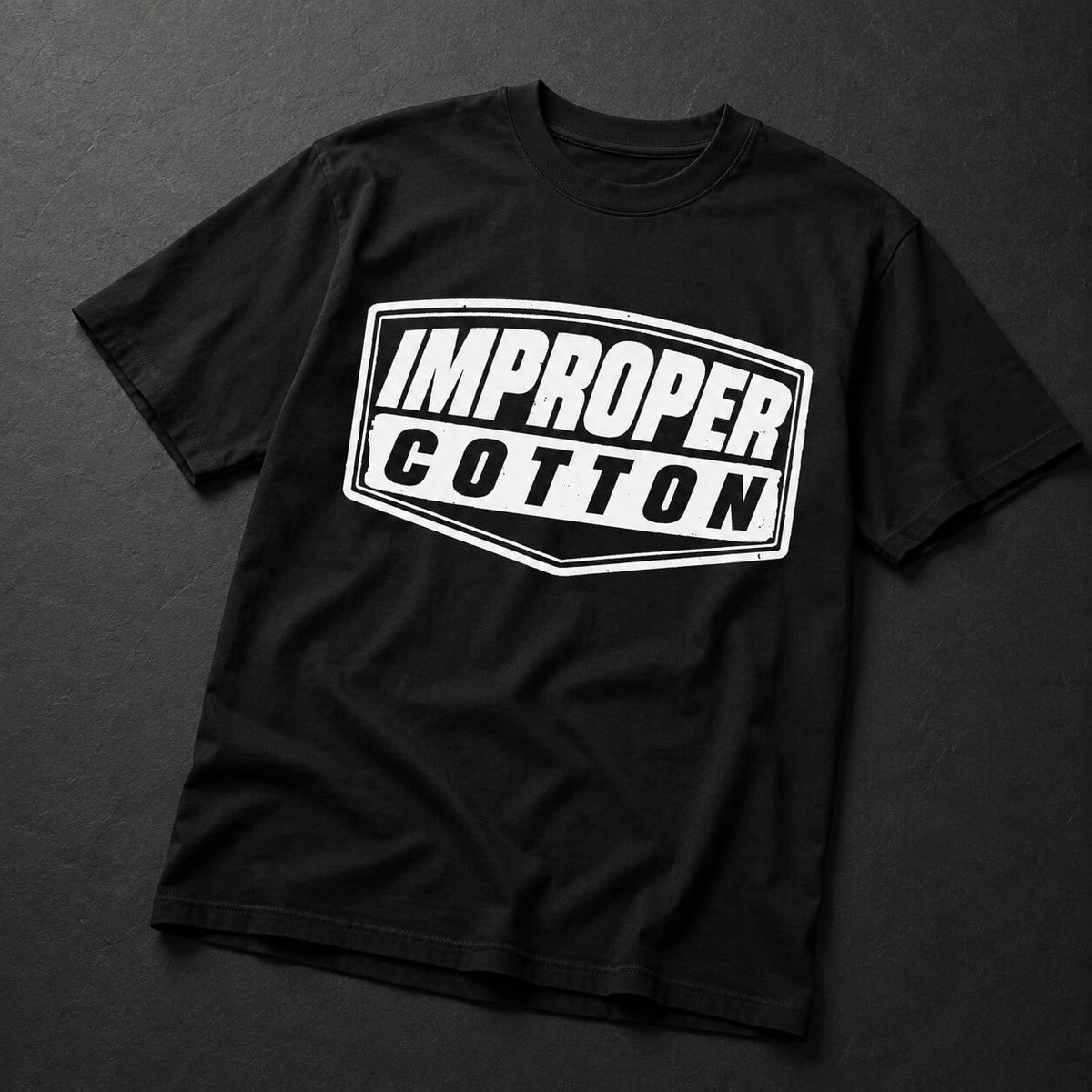 A black short-sleeve t-shirt with a white graphic print on the front that reads 'IMPROPER COTTON' inside a shield-like border, laid flat on a dark textured background.
